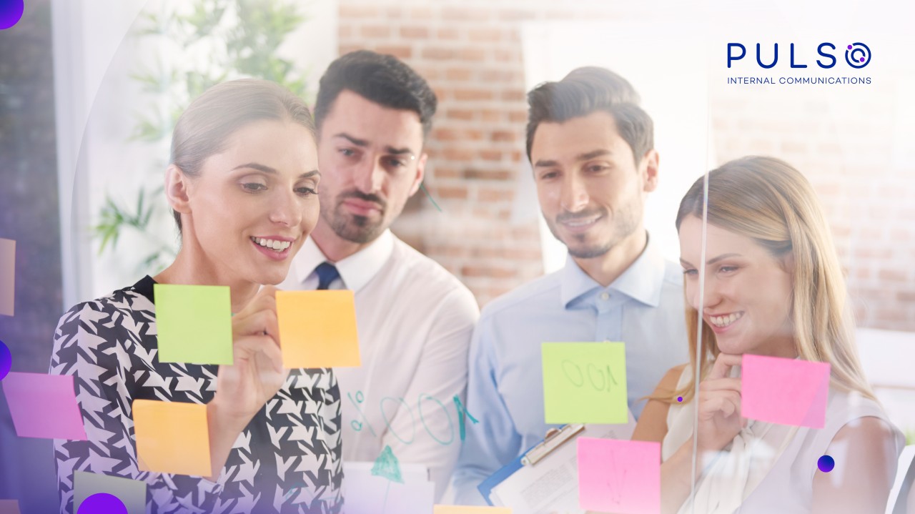 Tres profesionales sonrientes analizando notas adhesivas de colores en una pizarra de vidrio para planificar estrategias de comunicación interna y retención de talento.