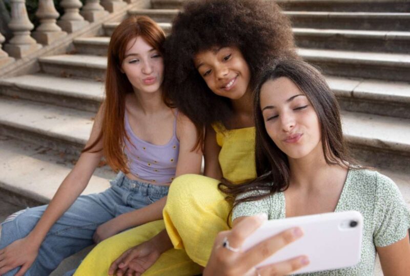 Tres mujeres jóvenes de la generación Z sentadas juntas al aire libre en unas escaleras mirando una pantalla de teléfono celular.