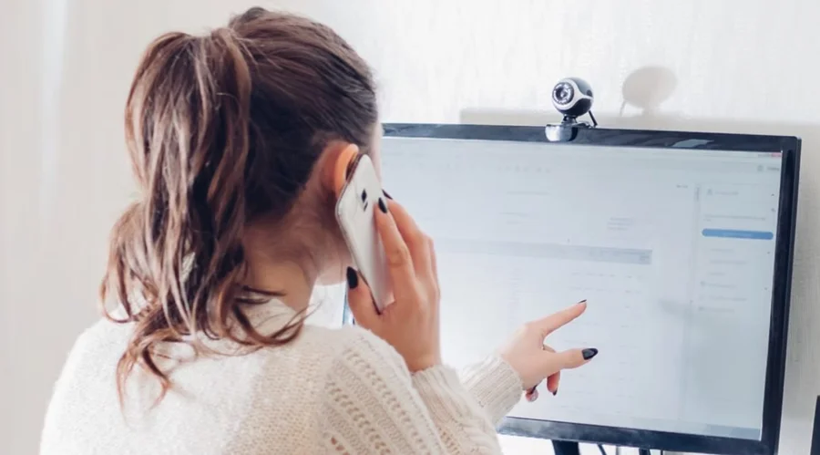 Mujer joven de espaldas hablando por teléfono celular frente al monitor de su computadora mientras trabaja en la oficina o hace home office.